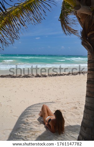 Brown skin girl in bikini lying on Mayan riviera coast on the sunny white sandy beach with coconut palm trees leaf admiring azure ocean after reopening because coronavirus covid-19 pandemic Mexico