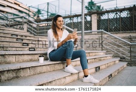 Young happy female in eyeglasses and casual clothes messaging and listening to music on mobile phone while sitting on outdoor stairs