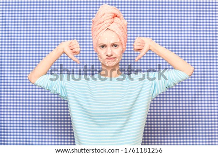 Portrait of unhappy sad girl with anti-acne skincare product on face, with towel on head, showing thumbs down sign of dislike, over shower curtain background. Care for skin and hair. Beauty concept