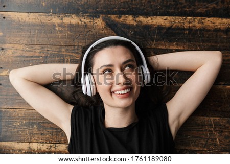 Photo from above of beautiful young brunette woman wearing headphones smiling and lying on wooden floor