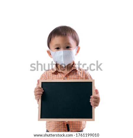 An Asian kid is standing hold a board with spaces for words.  And wear a surgical mask.