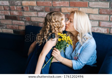 Daughter and mother are kissing in their home, girl gives mother a bouquet of flowers in cozy living room. Concept of Happy Mothers Day.