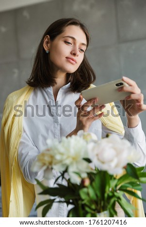 Beautiful lovely brunette woman taking a picture of a flower bouquet while standing at home