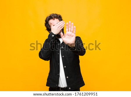 young crazy handsome man covering face with hand and putting other hand up front to stop camera, refusing photos or pictures against orange wall