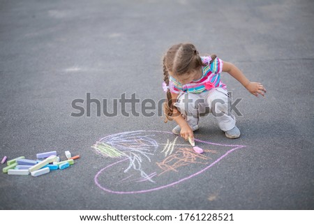Beautiful little girl draws heart with colorful crayons on asphalt, active games and leisure, street children’s entertainment.
