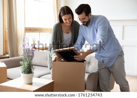 Happy young couple unpacking belongings, moving into new first house together, smiling husband holding frame with photo, family sharing good memories, relocation and mortgage concept