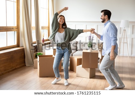 Happy young wife and husband dancing in modern living room with cardboard boxes with belongings, excited family celebrating moving day, satisfied customers relocating into new apartment, mortgage