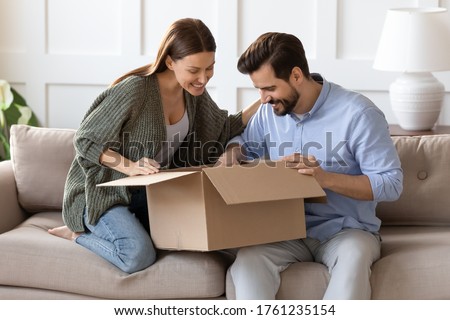 Happy curious couple unpacking parcel at home, sitting on couch, smiling woman looking into open cardboard box, satisfied clients customers received online store order, good delivery service Royalty-Free Stock Photo #1761235154