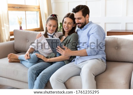 Happy parents with adorable daughter using computer tablet together, sitting on cozy couch in living room, smiling mother, father and little girl looking at screen, shopping online, enjoying weekend