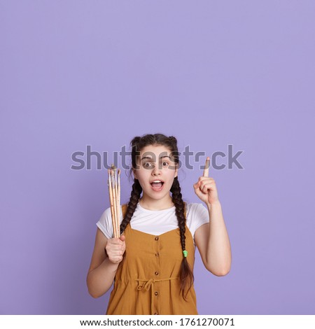 Surprised young girl artist with brushed in hands, pointing up with her index finger, keeps mouth opened, wearing casually, stands against lilac wall.