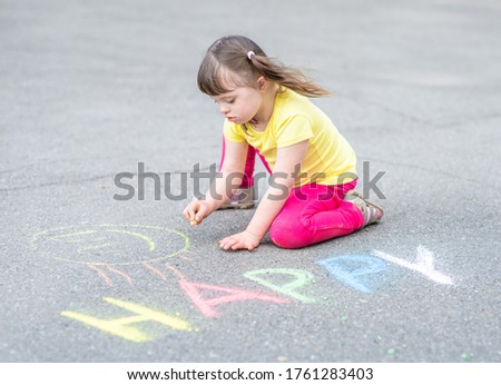 Little girl with syndrome down draws with chalk on the asphalt on summer day