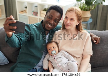 High angle portrait of young interracial family taking selfie photo with cute mixed-race baby while sitting on couch in home interior
