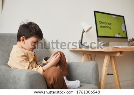 Side view portrait of cute boy sitting in armchair and using digital tablet with online school website in background, copy space