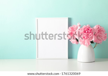 Home decoration with floral decor, frame poster on table. Beautiful flowers pink peonies in vase on mint background