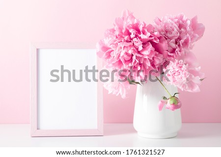 Home decoration with floral decor, frame poster on table. Beautiful flowers pink peonies in vase on pink background