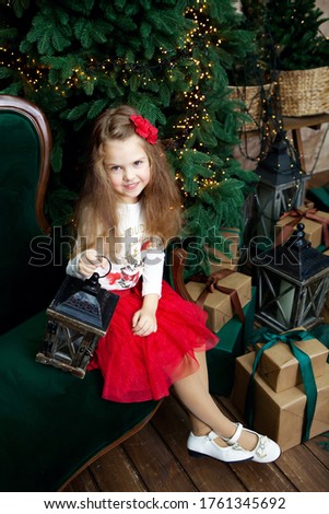 Cute girl in red dress sitting on sofa with candlestick on Christmas tree and gifts background. New year decorations