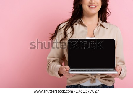 Cropped view of smiling woman showing laptop with blank screen on pink background