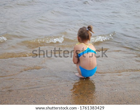 One  little girl, wearing blue swimsuit is sitting on  hunkers on the  beach during cold summer  vacation.