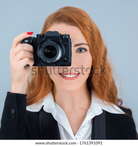 Young redhead woman holding a camera. She is wearing a suit.