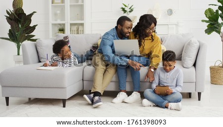 Happy african american family sitting on sofa together with device at home. Mother and father using laptop, small children brother and sister drawing at home, family time concept
