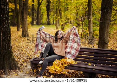 A girl with dark hair sits on a bench in the park in autumn and takes cover with a beautiful warm plaid . High quality photo