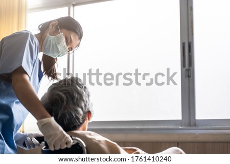 Asian nurse taking care of mature male patient sitting on wheelchair in hospital. Young woman and old man wearing surgical face mask for protection of covid 19 pandemic. Girl smile to elderly man.