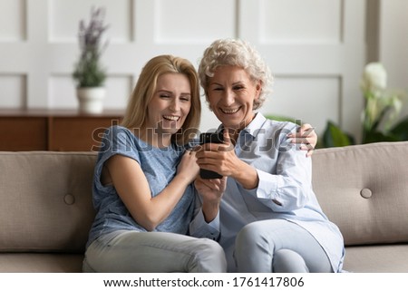 Funny elderly grandma and teen adult granddaughter having fun seated on couch using smartphone cool application. Aged mother grown up make videocall chatting with relatives by videoconference concept