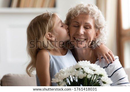 Close up image happy old grandmother closed eyes enjoy moment while her little granddaughter kisses on cheek and congratulates her with birthday presents flowers. Women 8-march Day, life event concept