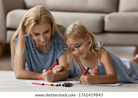 Young mother lying on warm floor with little daughter, parent helps to preschool kid girl, teaches her to draw on album paper using colored felt-tip pens enjoy hobby pastime activity at home concept