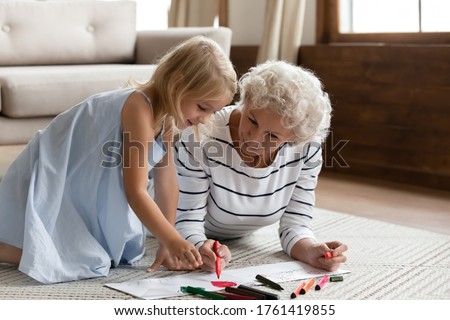 Elderly babysitter teach little girl drawing lying on warm floor, helping improve creative abilities, develop artist talent. Caring old 50s grandma spend time with small granddaughter at modern home
