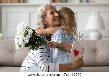 Sincere old grandma hugs small attentive granddaughter express her gratitude for received surprise at birthday or 8-march Woman Day. Grandchild makes surprise for granny gave her flowers and postcard Royalty-Free Stock Photo #1761419900