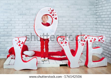 Little girl standing in front of decoration for St. Valentine's day with big letter O, grey brick wall as a background