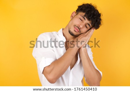 Relax and sleep time. Tired young handsome man with closed eyes leaning on palms as pillow pretending sleeping being exhausted seeing dreams standing against yellow background. Sleeping gesture.