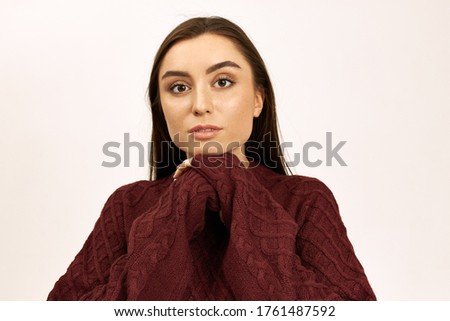 Portrait of beautiful stylish young female with brown eyes and straight hair posing isolated with hands clasped wearing oversize long sleeved pullover, being cold on winter day. Season and clothing