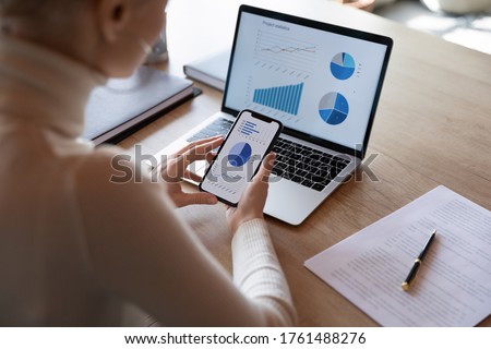 Woman hold smartphone use pc at workplace. Project stats financial data sales charts on laptop and cellphone screen, close up view over shoulder. Report preparation, synchronization for safety concept Royalty-Free Stock Photo #1761488276