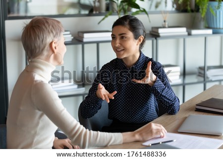Indian female employee talking with Caucasian mate seated at workplace desk expresses her opinion on current issue, proposes solution to problem, share thoughts while working on common project concept Royalty-Free Stock Photo #1761488336