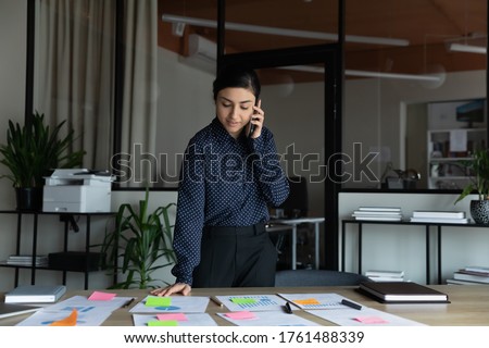 Indian businesswoman project leader prepare presentation for company investors use colorful post-it notes printed graphs charts financial data overview and forecast talk by phone standing at boardroom