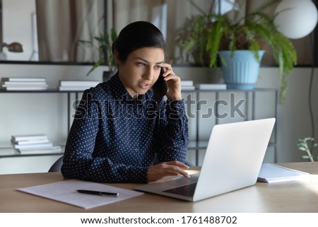 Indian ethnicity businesswoman search on laptop information while talking with client on phone. Manager agent solve help to company customer distantly, modern tech usage, busy fruitful workday concept Royalty-Free Stock Photo #1761488702