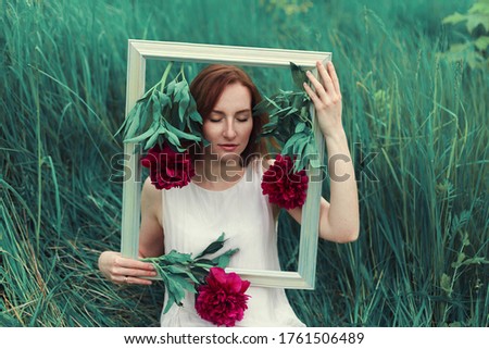 Redhead girl with closed eyes holds picture frame decorated with flowers in front of her