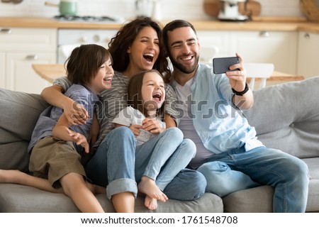 Close up happy parents with little children using smartphone together sitting on couch at home. Smiling father holding phone taking selfie with kids. Family watching video having fun with cellphone.