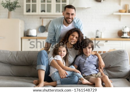 Close up portrait picture of happy young family of four people sitting on couch at home. Smiling parents with little children hugging looking at camera posing for photo in living room.