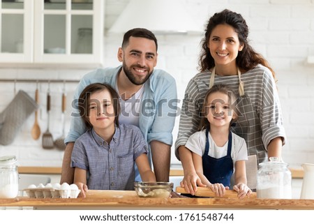 Close up headshot portrait picture of happy young couple with children cooking dinner looking at camera standing at table in kitchen. Smiling family using rolling pin for pastry on wooden board.