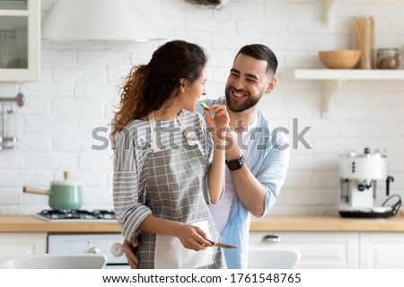 Close up happy couple playfully cooking cooking salad in kitchen. Hugging smiling husband looking at wife eating fresh cucumber preparing dinner or breakfast. Family enjoying weekend together.