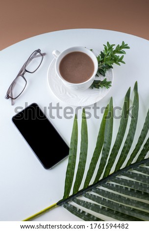 Smart phone, chocolate coffee, white cup, glasses, green decor plant, palm leaf on white table, top view.