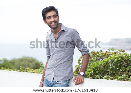 Happy handsome man standing outdoors smiling happy, laughing joufully with green bush tree blue sky and sea on background