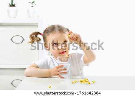 a child looks at a capsule with fish oil, tastes omega3, a glass of water in his hand and a capsule, is in a bright room, takes pills, yellow capsules, white table, white background, copy space Royalty-Free Stock Photo #1761642884