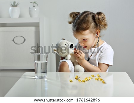 A glass of water and fish oil pills. A child plays doctor in the children's room, a scattering of yellow capsules on the table, a bear cub takes omega 3 pills. a Child with a stethoscope in his ears. Royalty-Free Stock Photo #1761642896
