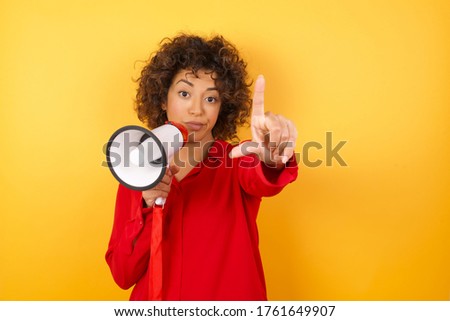 young arab woman wearing red shirt holding a megaphone over yellow background making fun of people with fingers on forehead doing loser gesture mocking and insulting.