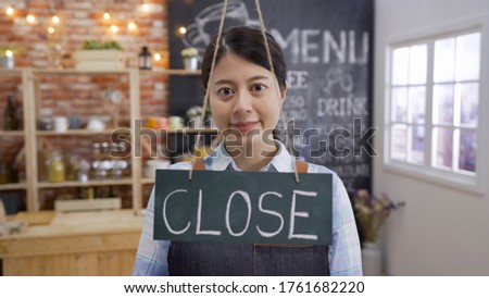 close sign. Joyful positive woman smiling while putting door plate on window in cafe bar. asian female waitress worker looking camera cheerful elegant face wear apron work in coffee shop indoors.