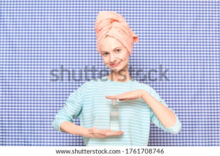 Portrait of cute happy blond girl with anti-acne skincare product on face, with towel on head, smiling, holding cosmetic bottle, over shower curtain background. Care for skin and hair. Beauty concept